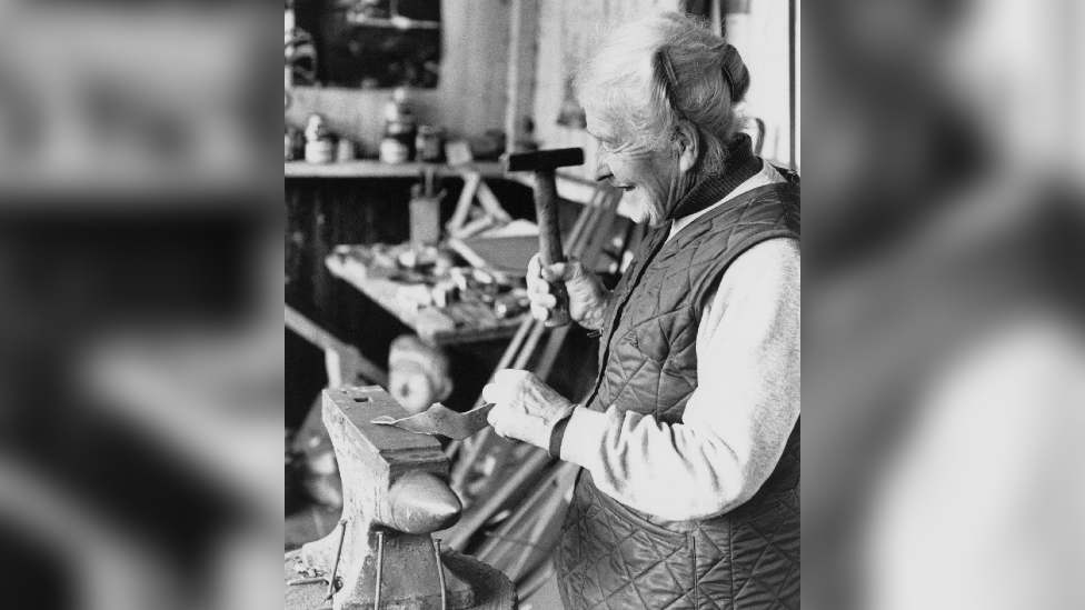 An elderly woman holding a hammer, hammering a piece of metal on an anvil