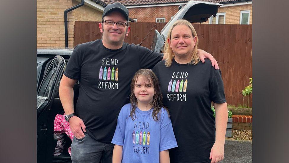 Rosie, in the front and centre frame, with her dad James and mum Julie standing behind her. They are all standing closely together, and James has his arm around Julie's shoulders. They are smiling and standing outside, in front of a dark coloured car that has a rear door and boot open. A garden fence and housing can be seen in the background. Rosie is wearing a purple T-shirt with "SEND Reform" written on it. James and Julie are wearing black T-shirts with the same slogan written on them.