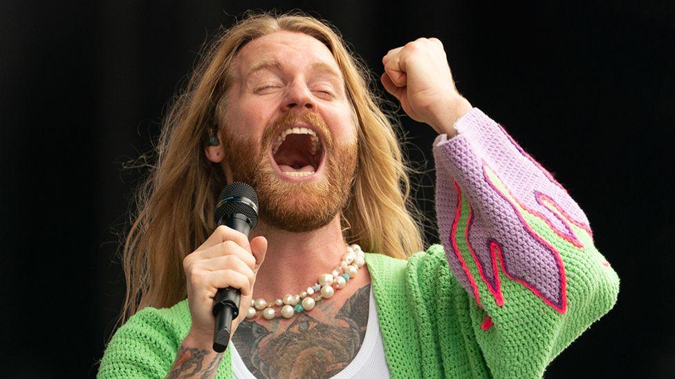 Sam Ryder performing on stage. He is singing with his eyes closed, his mouth wide and is holding his fist in the air. He has very long fair hair, a beard and is wearing a pearl necklace and green knitted cardigan with purple flames on the end of the sleeves and has a tattooed chest