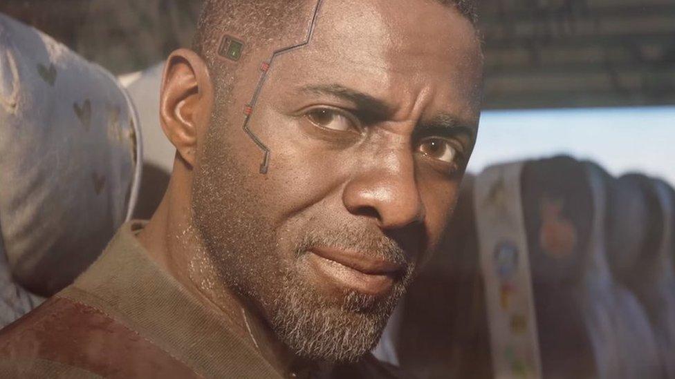 A computer-generated Idris Elba in close-up, sitting in the window seat of a train carriage. His short, stubbly beard is illuminated by sunlight from outside and he has a serious, thoughtful expression. Some sort of wire/track runs from his temple to his cheek, giving him a futuristic appearance.