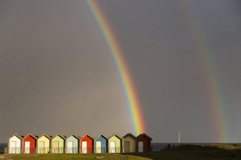 A rainbow arcs across a dark sky and down on to a row of colourful beach huts