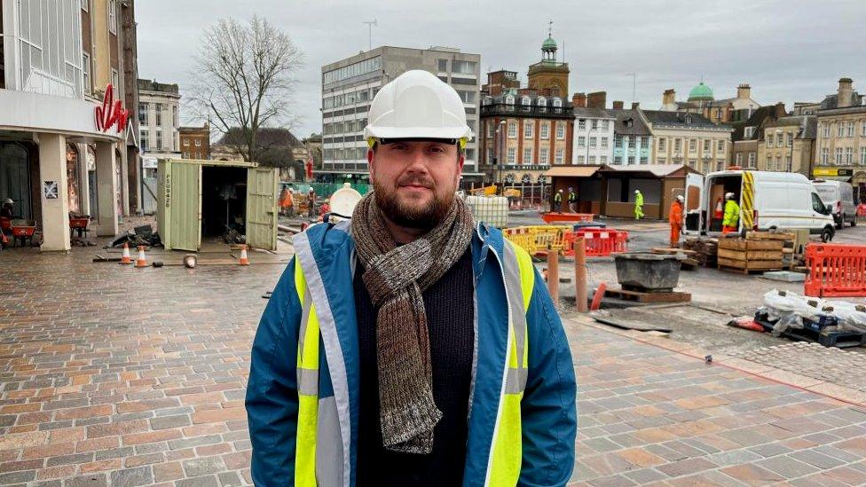 Bearded man in blue jacket and yellow hi-viz stands in market square