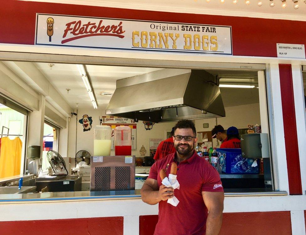 A man standing outside a corn-dog stand holds two corn dogs in his hands