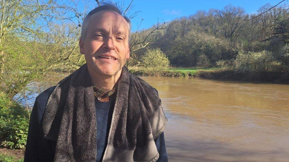 A man standing by a river with his towel round his neck