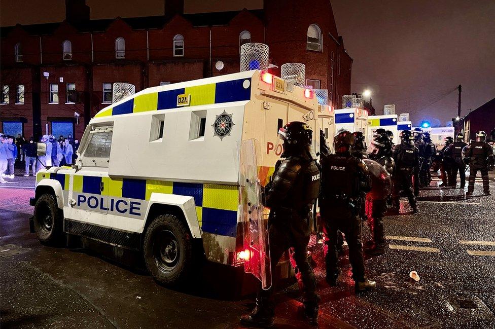 Police officers in riot gear stand behind armoured police Land Rovers