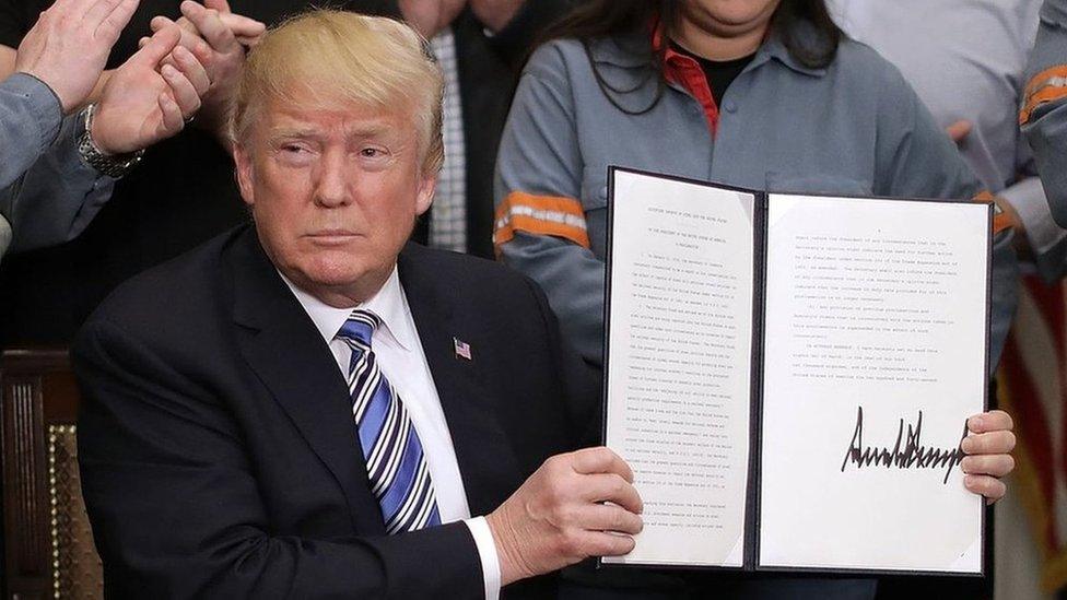 President Donald Trump holds up a piece of paper with his signature while surrounded by steel workers in uniform