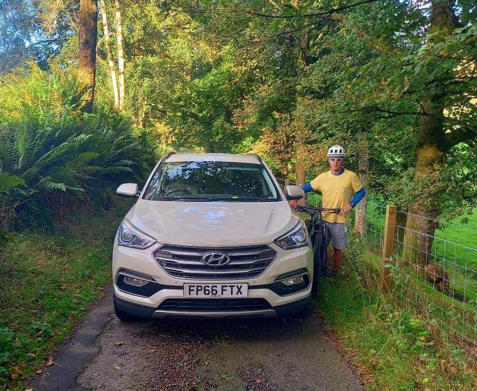 Narrow road with a car filling the entire width. A cyclist is squashed at the side.