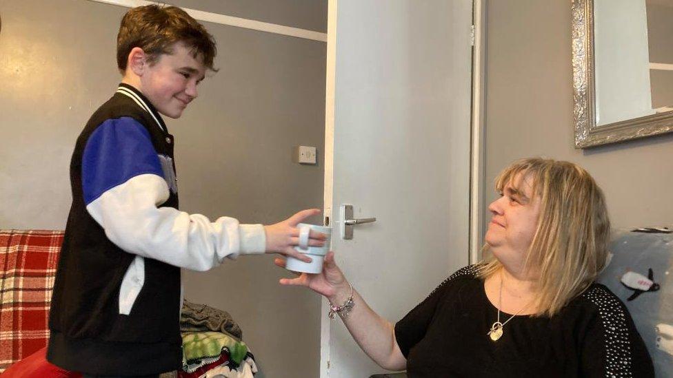 Boy wearing blue and white handing a mug to his mother who is sitting on a sofa