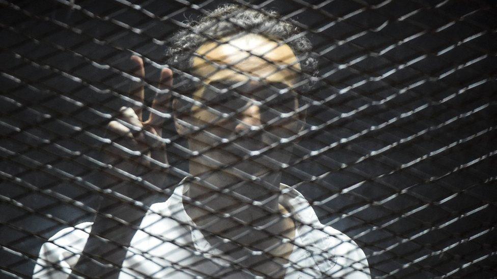 Egyptian photographer Mahmoud Abdel Shakour Abou Zeid, also known as Shawkan, flashes the victory gesture from inside a soundproof glass dock, during his trial in the capital Cairo on July 28, 2018