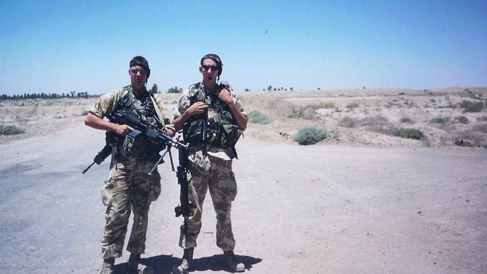 Two men in army uniforms stand holding machine guns in an empty desert/field