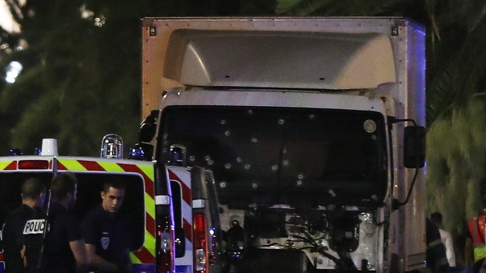 Lorry, showing its windscreen full of bullet holes, with a police van and police officers in front