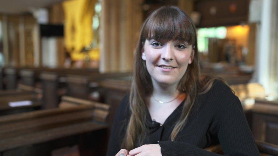 Elise, with long dark hair, sits in a church where a concert is taking place