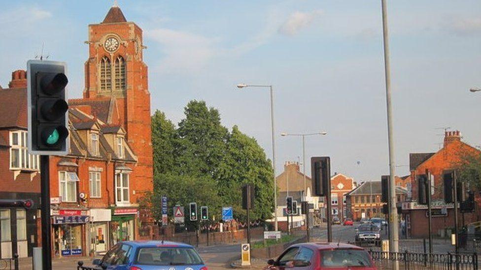 St James Road - showing a brick-built church to the left with a tower, and shops to the right. There are traffic lights and a pedestrian crossing across both sides of the carriageway.  Some cars and a van are visible on the road.