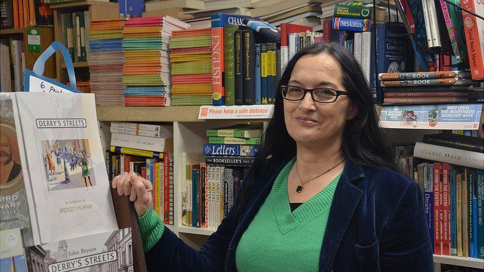 Jenni Doherty is wearing glasses, a velvet blazer and green jumper and is standing in front of a shelf of books 