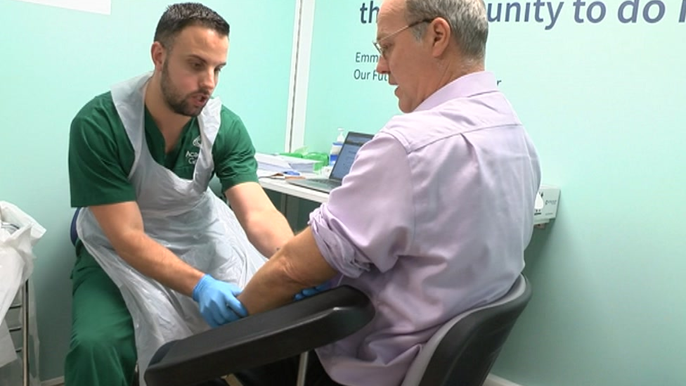 A man in scrubs and a uniform holds the arm of a man with his sleeve rolled up