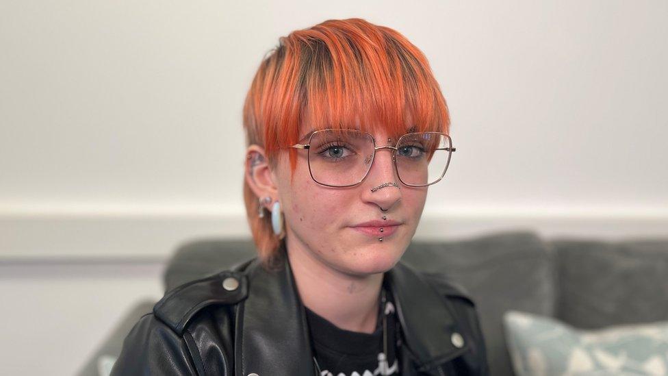 Caragh, a young woman with short hair dyed orange, square glasses and piercings