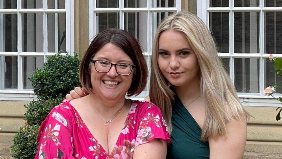 Angharad with her mum Andrea posing for the camera, both are smiling. Andrea is on the left and has glasses and a short brunette bob hairstyle. She is wearing a flowery pink top and has a silver necklace around her neck. Angharad is to her right and has her right arm around her mum's shoulder. She has medium length blonde hair and is wearing a teal dress that is just over her right shoulder.