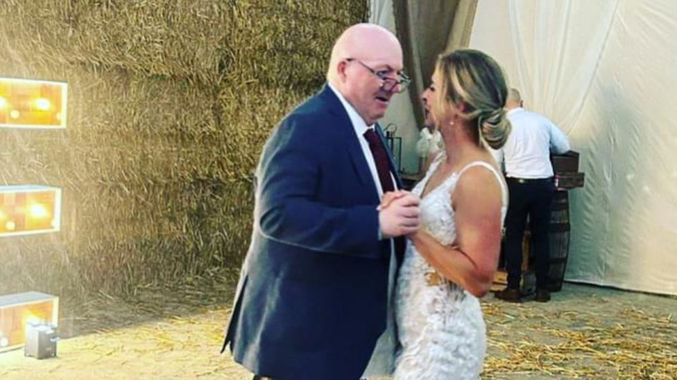 Roger in a suit and Jessie in her wedding dress dancing in front of decorations and hay bails