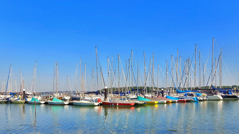 Blue skies over a marina with lots of colourful sail boats moored up in clear waters.