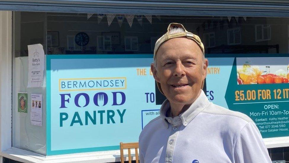 Ken Blower wearing a white t-shirt and a cap on backwards stands in front of a shop front that says food pantry