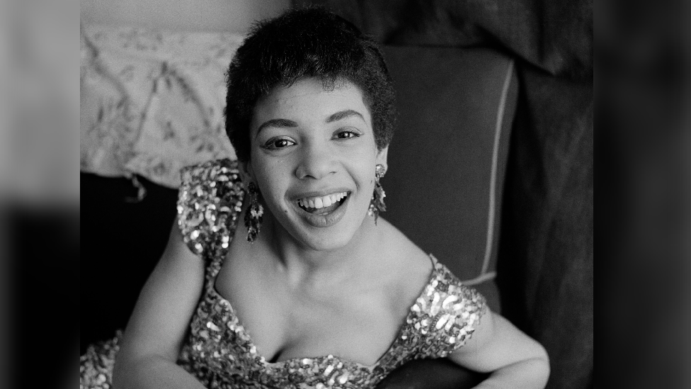 A black and white photo of young Shirley Bassey wearing a sequined dress in 1956. She is looking at the camera