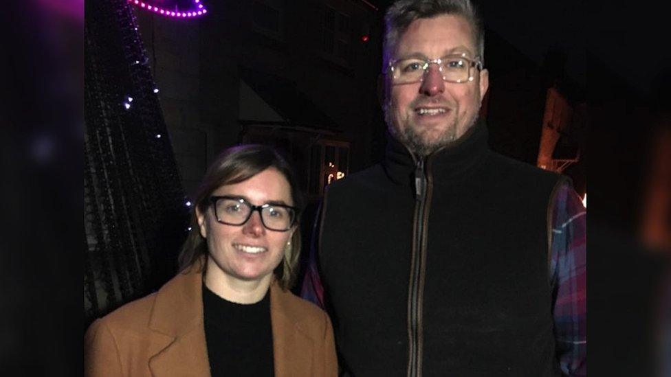 Faye and Richard. She is on the left, wearing glasses, a black top and a brown jacket. Richard is stood next to her on the right, wearing a black gilet and check shirt. They are both looking directly at the camera and smiling.