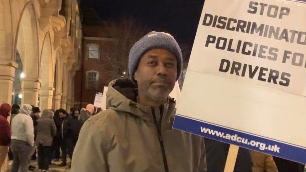 Man in blue woolly hat and anorak carrying banner saying "stop discriminatory policies for drivers"