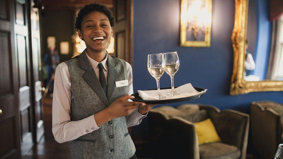 A smiling waitress in a waistcoat holds a tray with two glasses of white wine on