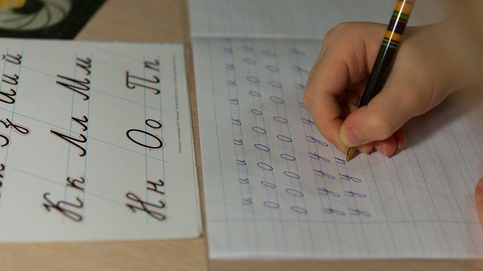 A child copies letters in joined style from a workbook, their page showing line after line of the letters u, o, and y