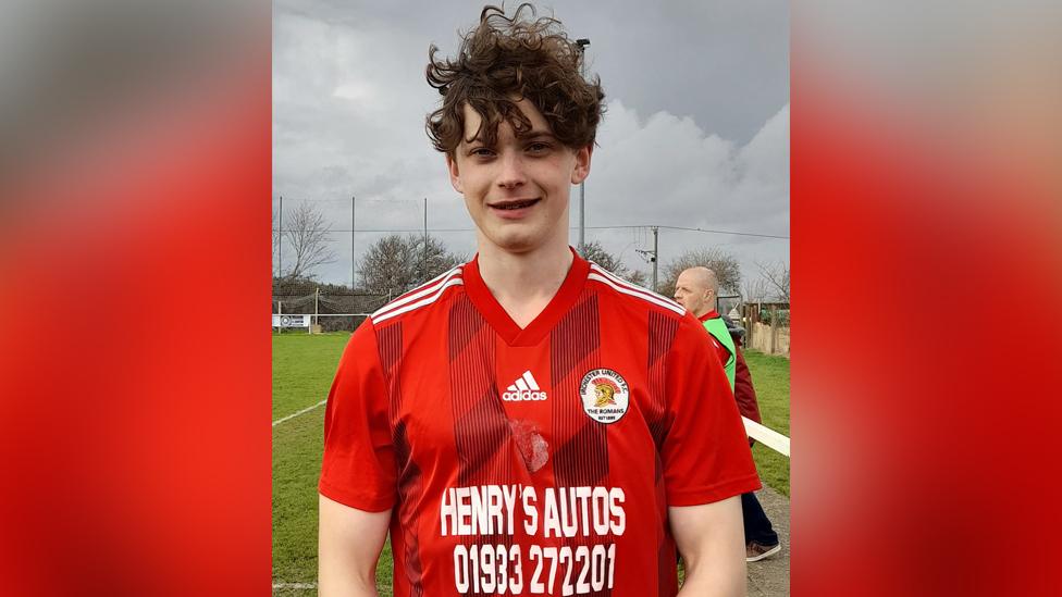 Brandon Thacker wearing Irchester Romans kit; a short sleeved red shirt with a v neck collar