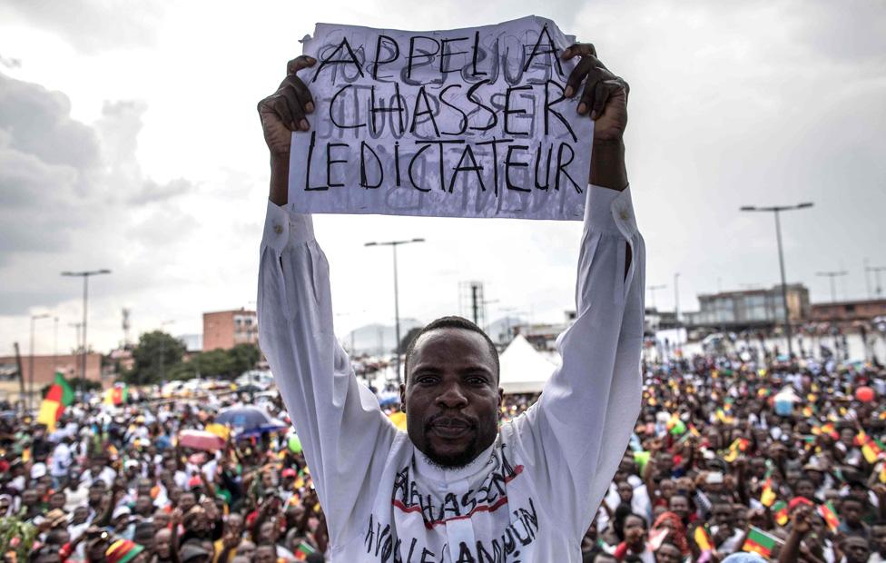 A man holds a paper with the writing in French "A call to dismiss the dictator" in front of a crowd of supporters of the leader of the Cameroonian opposition party Movement for the Rebirth of Cameroon (MRC) during an electoral campaign rally on September 30, 2018