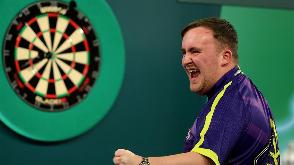 A darts player celebrates, with a dart board in the background. He wears a purlpe and yellow short-sleeved T-shirt