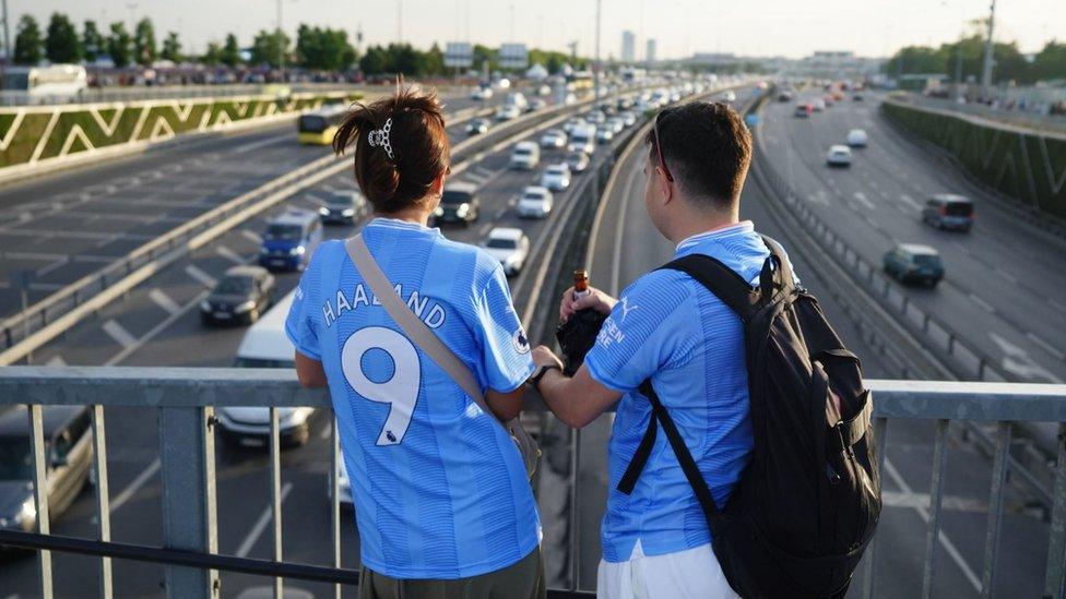 Manchester City fans stood on motorway bridge looking at queue of traffic