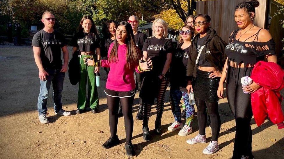 Sheetal wearing casual spots clothes and a punk hoodie stood in front of a group if performers presenting to another camera