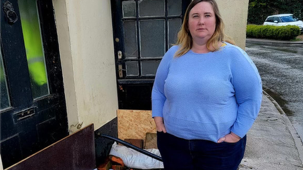 Woman stands in front of flood barrier with a muddy road behind her