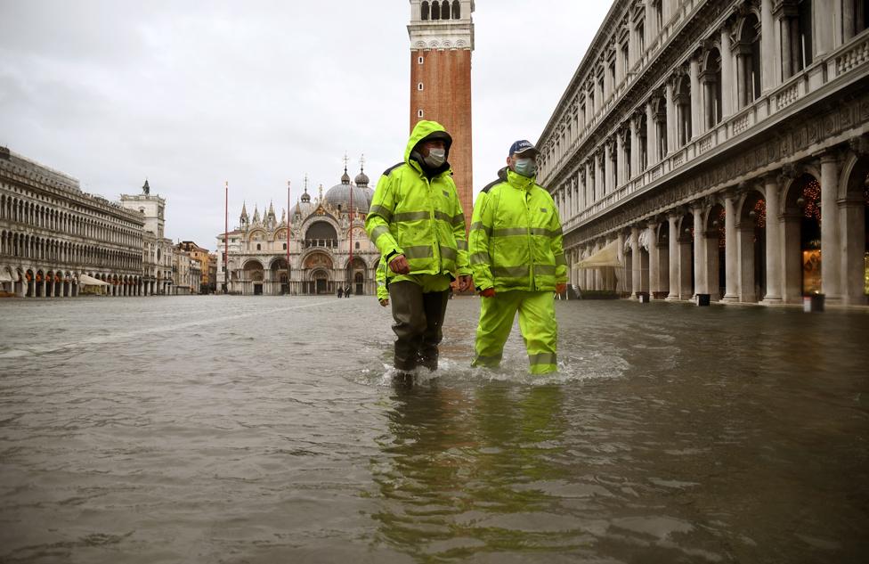 Men wearing waterproof gear walk through floodwater in St Mark's Square, Venice, Italy