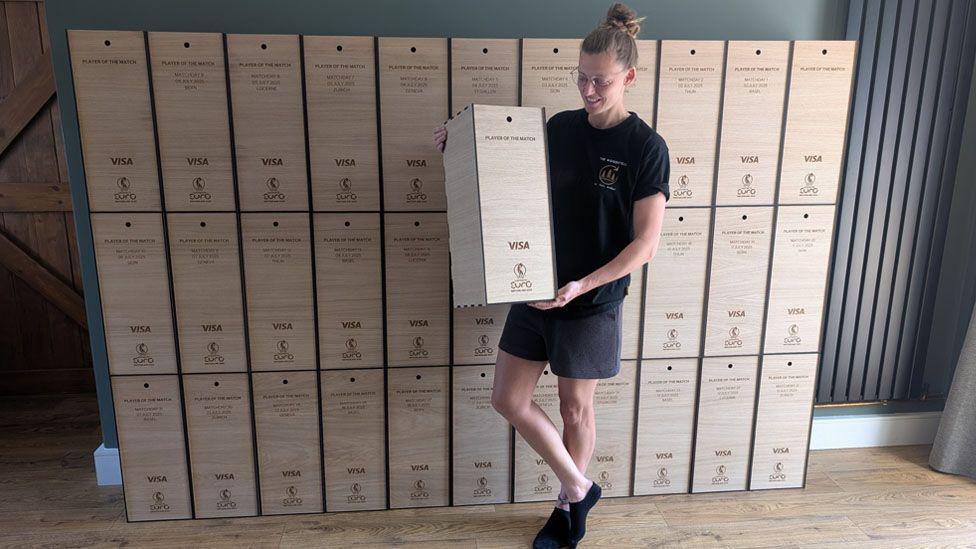 Yana is wearing black t-shirt and dark grey shorts and black socks. She is standing holding gone of the presentation boxes in front of a number of boxes stacked up in a row behind.
