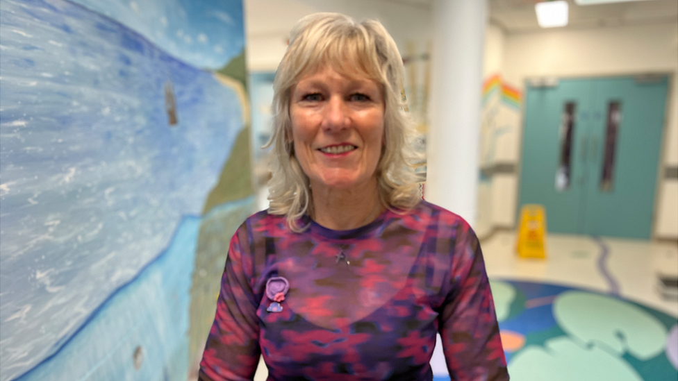 A woman in a pink top with blonde hair standing in a hospital ward