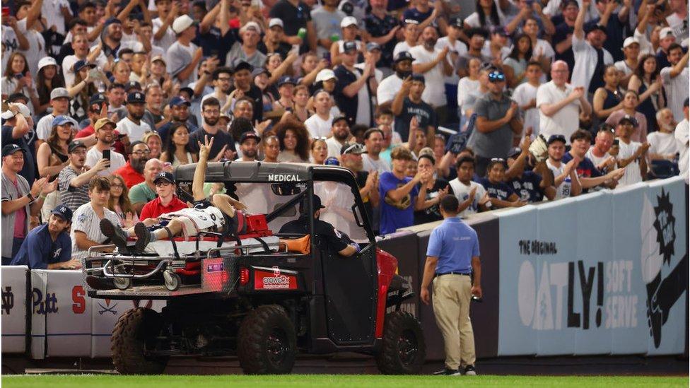 The cameraman holding up a hand to wave as he is being taken off the field