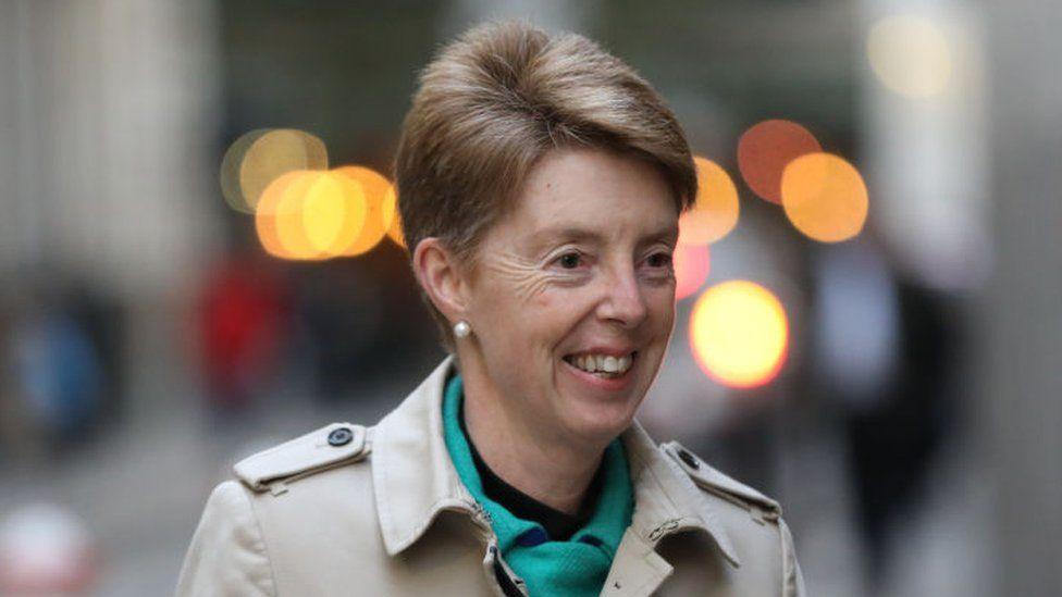 A smiling Paula Vennells is pictured on a busy street. She wears a beige raincoat and a green scarf.