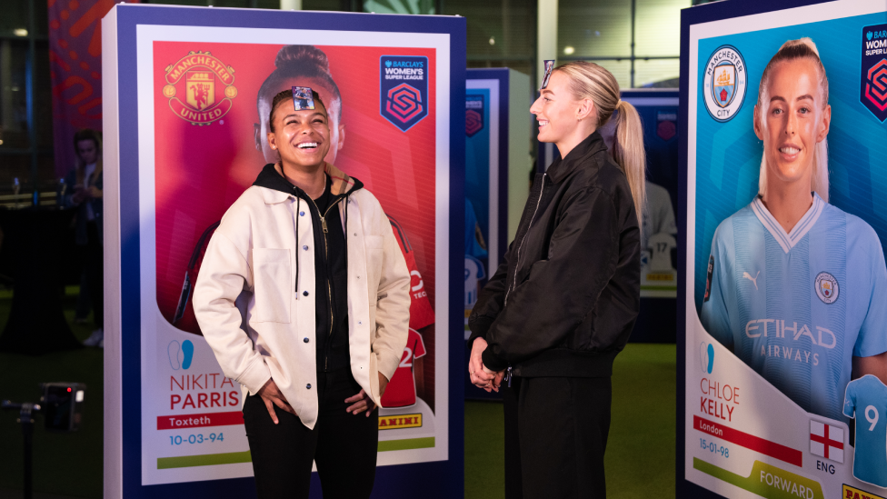 Chloe Kelly and Nikita Parris standing in front of boards which show their life-size Panini stickers. Both have stickers displaying their pictures stuck to their foreheads, and are laughing and smiling. Nikita in particular, sports a big beaming smile. Chloe's standing side-on to the viewer and smiling as she looks at Nikita.