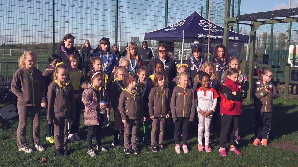 About 20 girls in brownie and guide uniforms standing in rows in front of the nets at a training ground