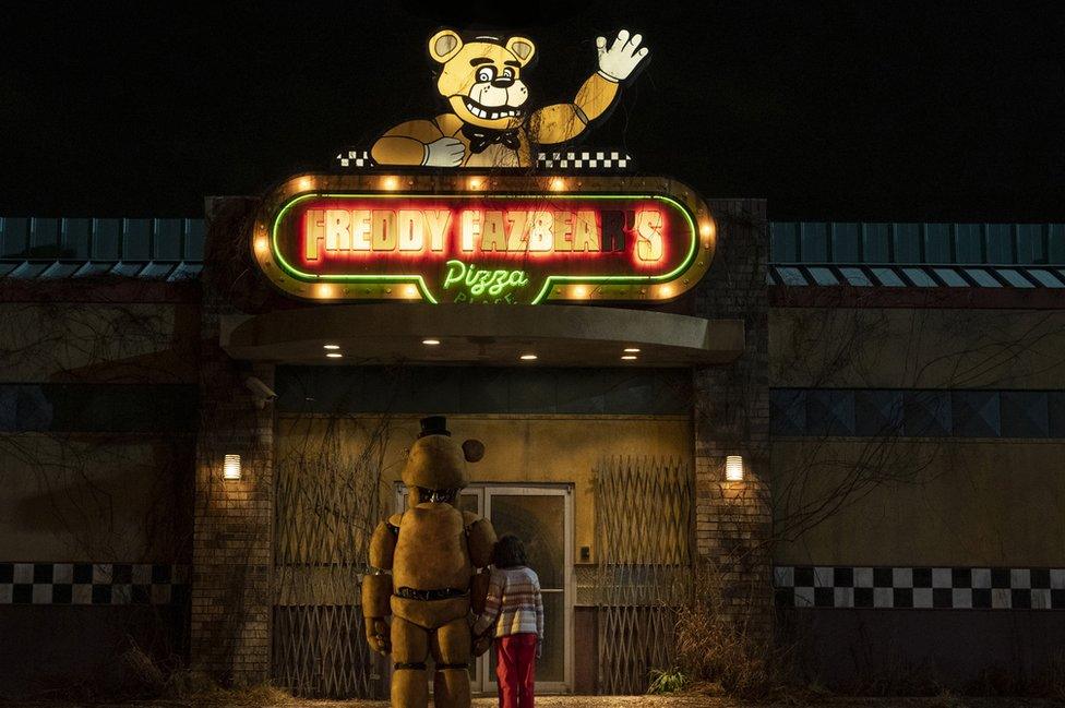 A fast food restaurant, shrouded mostly in darkness, with a slightly grubby neon sign above that reads "Freddy Fazbear's Pizza" in block capital letters. Above the lettering is an illuminated bear character. He's smiling and has one arm raised in a wave. In the foreground, a small child and a mechanical bear face away from the camera, holding hands.