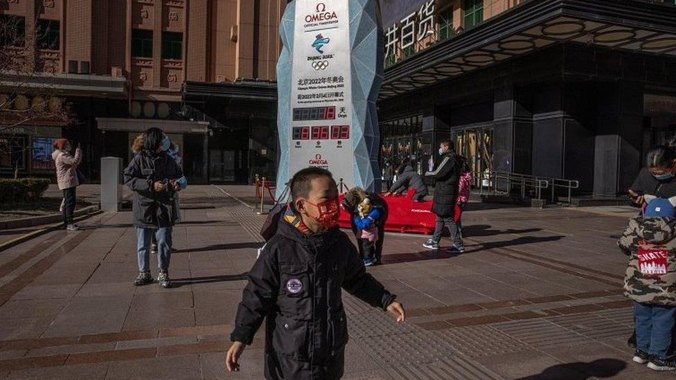 Parents with their children wearing face masks stand next to a countdown display showing days left for the Beijing 2022 Winter Olympics, on a shopping street in Beijing, China, on 17 January 2022