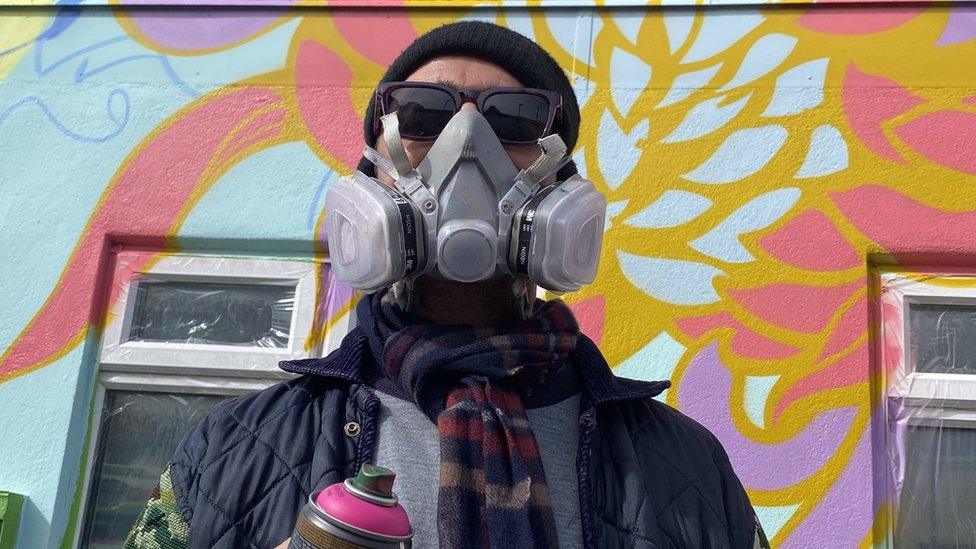 A street artist in a fume mask holding a can of pink spray paint in front of a wall spray painted with bright flowers