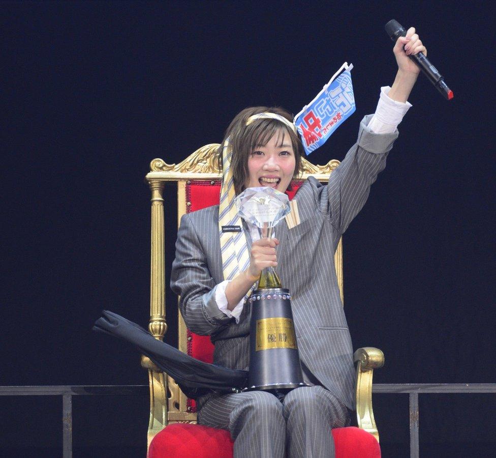 Miku Tanabe in a grey pin-stripe suit with a neck tie around her head, seated on a red throne-like chair and with a trophy on her lap.