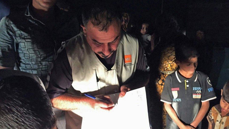 Charity worker writes on paper by limited electric light, surrounded by boys and men