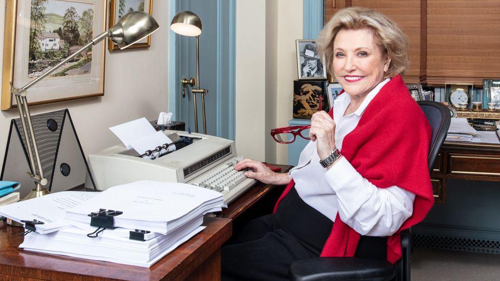 Barbara Taylor Bradford sitting at a typewriter in an office. she is wearing a white shirt and a red shawl and is holding her red glasses in her left hand.