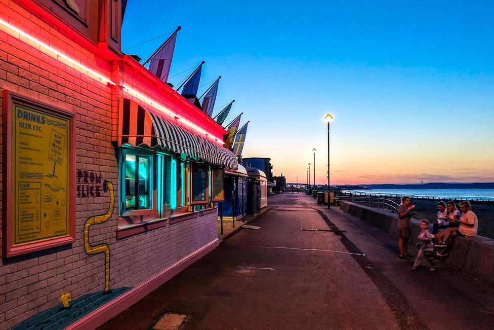 The Sea Is Too Hot - a group of people on a seaside promenade, next to a building illuminated by neon lights