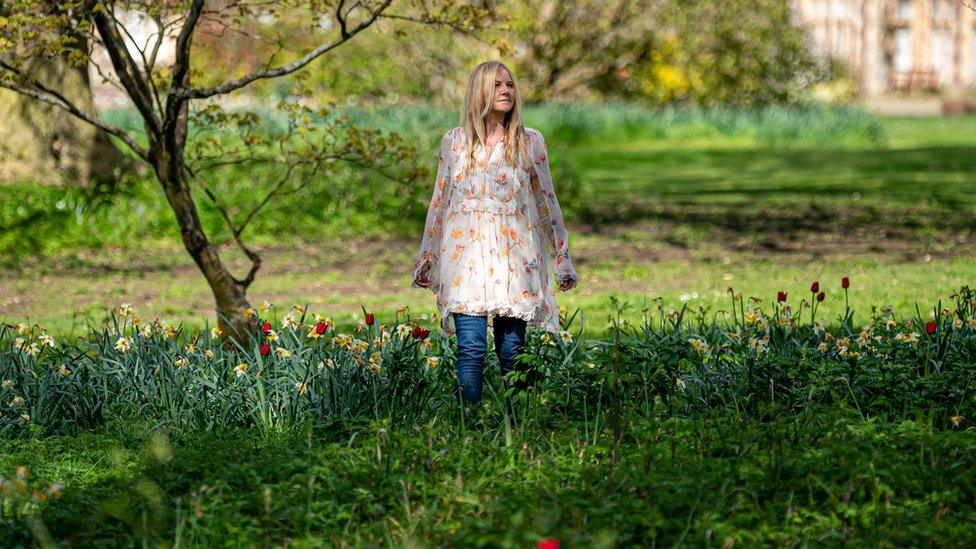 Sarah Class standing in a patch of flowers wearing a white floral dress and blue jeans